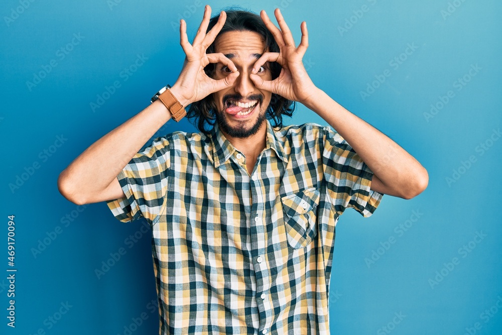 Young hispanic man wearing casual clothes doing ok gesture like binoculars sticking tongue out, eyes looking through fingers. crazy expression. Young hispanic man wearing casual clothes doing ok gesture like binoculars sticking tongue out, eyes looking through fingers. crazy expression.