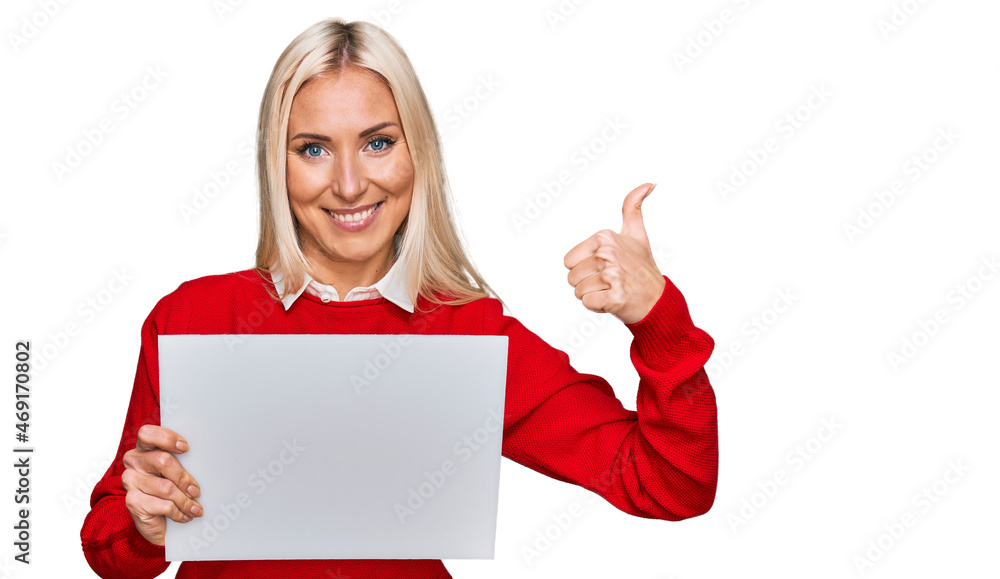 Young blonde woman holding blank empty banner smiling happy and positive, thumb up doing excellent and approval sign Young blonde woman holding blank empty banner smiling happy and positive, thumb up doing excellent and approval sign
