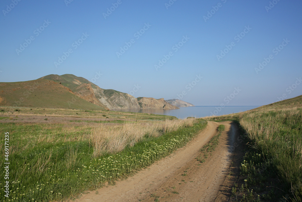 A road on the background of rocky mountains and the sea in the middle of blooming meadows