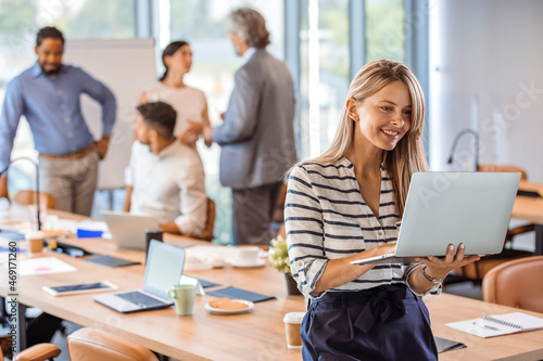 Woman manager holding laptop and standing in modern office. Businesswoman in casual wear holding laptop smiling and looking to camera while her colleagues communicating in the background