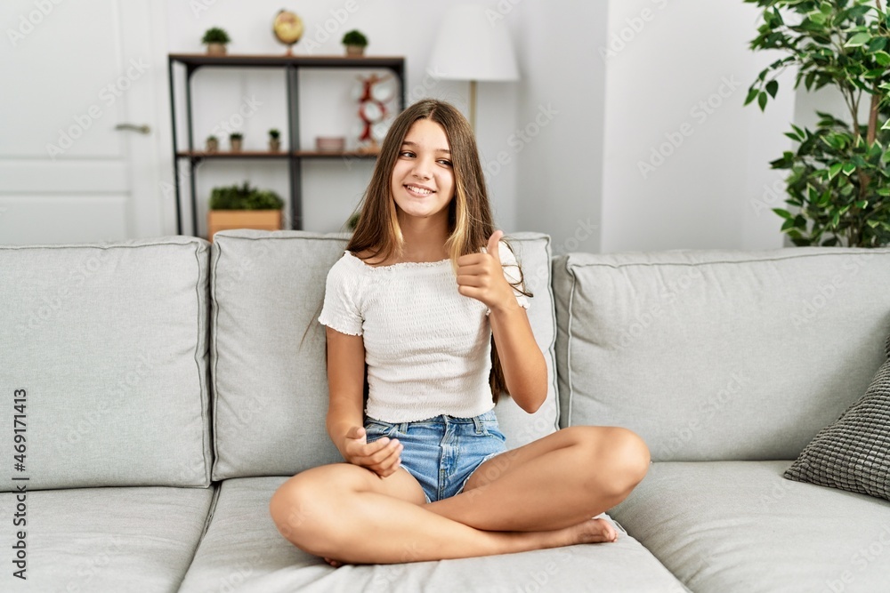 Young brunette teenager sitting on the sofa at home doing happy thumbs up gesture with hand. approving expression looking at the camera showing success.