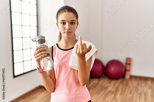 Young brunette teenager wearing sportswear holding water bottle showing middle finger, impolite and rude fuck off expression