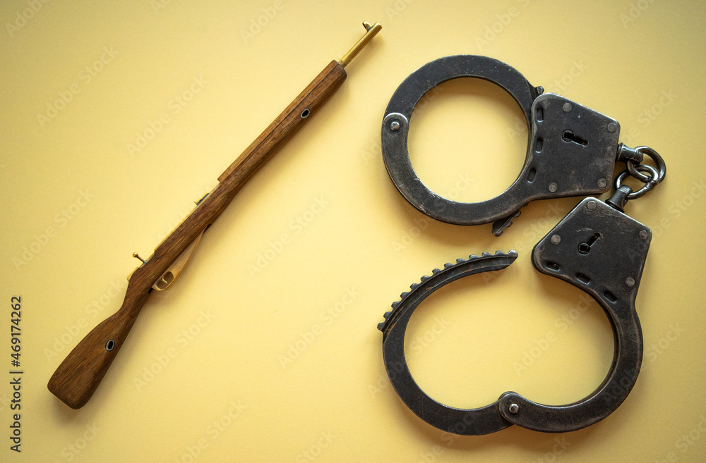 A wooden rifle on a yellow isolate with open metal handcuffs. The law on weapons. Manufacture and storage of firearms. Tightening of the law on weapons. Punishment for breaking the law. A wooden rifle on a yellow isolate with open metal handcuffs. The law on weapons. Manufacture and storage of firearms. Tightening of the law on weapons. Punishment for breaking the law.