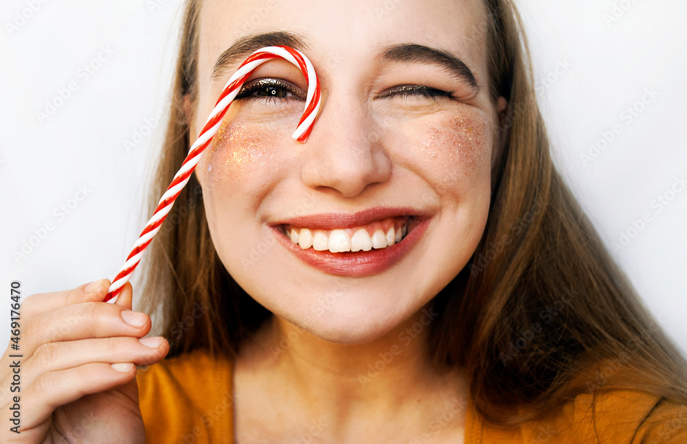 Close portrait of a girl with Christmas lollipops. Christmas candy. A woman with sparkles on her cheeks closes one eye against white. Close portrait of a girl with Christmas lollipops. Christmas candy. A woman with sparkles on her cheeks closes one eye against white.