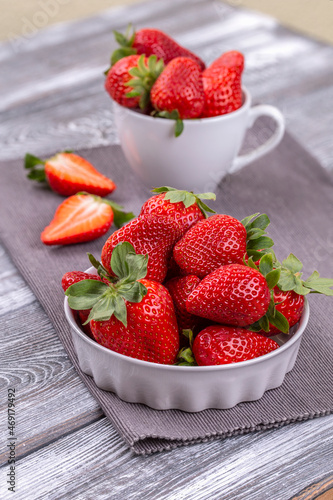 Juicy strawberries in a white ceramic bowl and cup, sliced berries on a gray shabby wooden table in a rustic style, copy space, vertical photo.