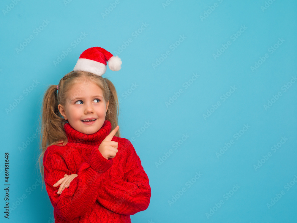 Happy surprised child in Santa hat and red sweater pointing to empty copy space on blue background. Happy surprised child in Santa hat and red sweater pointing to empty copy space on blue background.
