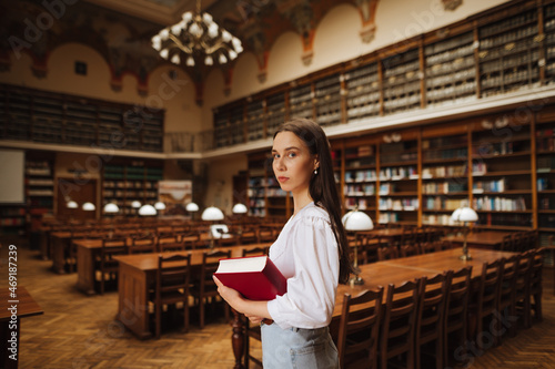 Portrait of attractive female student with book in hands at university library, looking at camera with serious face on background of reading tables.