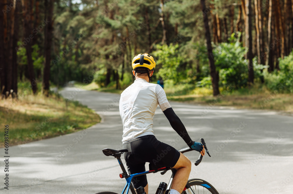 Professional cyclist trains in the woods in the fresh air, stands on an asphalt road with a beautiful view of the coniferous forest, back view.