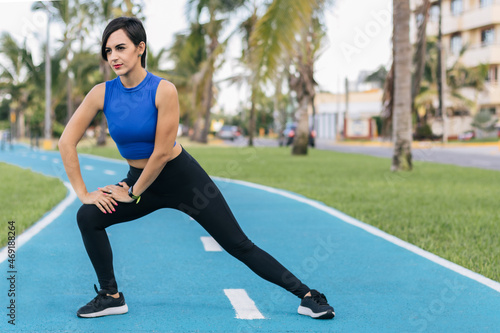 Hispanic young adult woman stretching her legs before doing exercise at a park leaning to the left