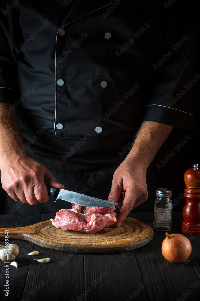 A professional chef cuts meat with a knife in the kitchen prepares food. Vegetables and spices on a table in a restaurant for preparing a delicious lunch