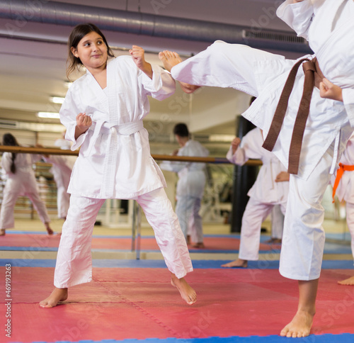 Young girl in white kimono blocking kick during sparring karate training in gym.