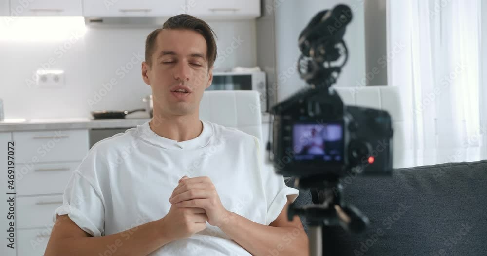 Millennial blogger or vlogger man filming new vlog video with professional camera, mounted on tripod sitting on sofa at home. Millennial blogger or vlogger man filming new vlog video with professional camera, mounted on tripod sitting on sofa at home.