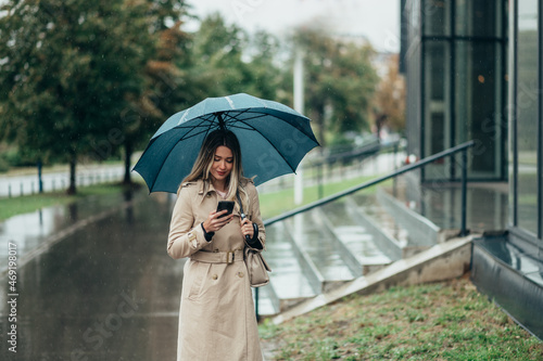 Businesswoman with umbrella using smartphone and walking down city street during rain