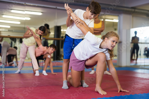 Sportive preteen kids practicing in pair self-protection in class with female teacher