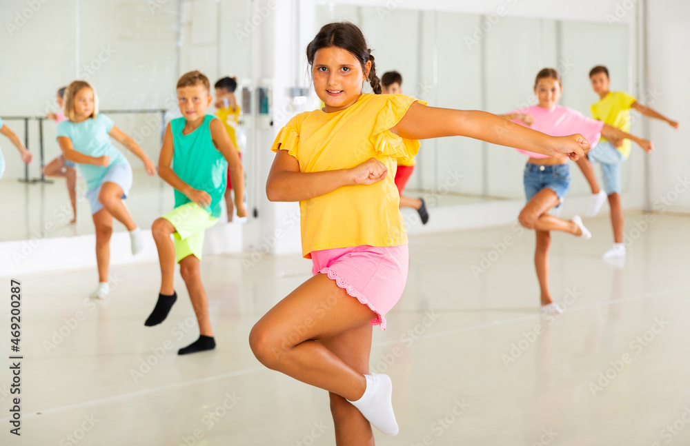 Smiling latin american preteen girl training movements of modern vigorous dance with group of tweens in children dance studio.