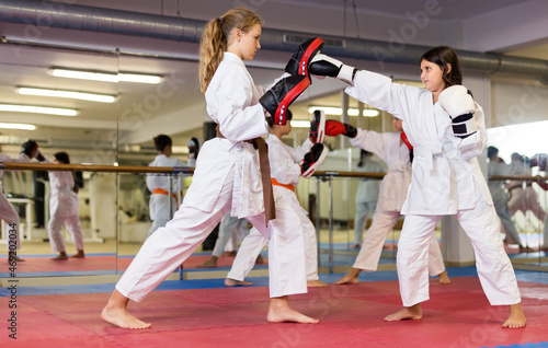 Two young girls in boxing gloves and focus mitts training punches during group karate training in gym.