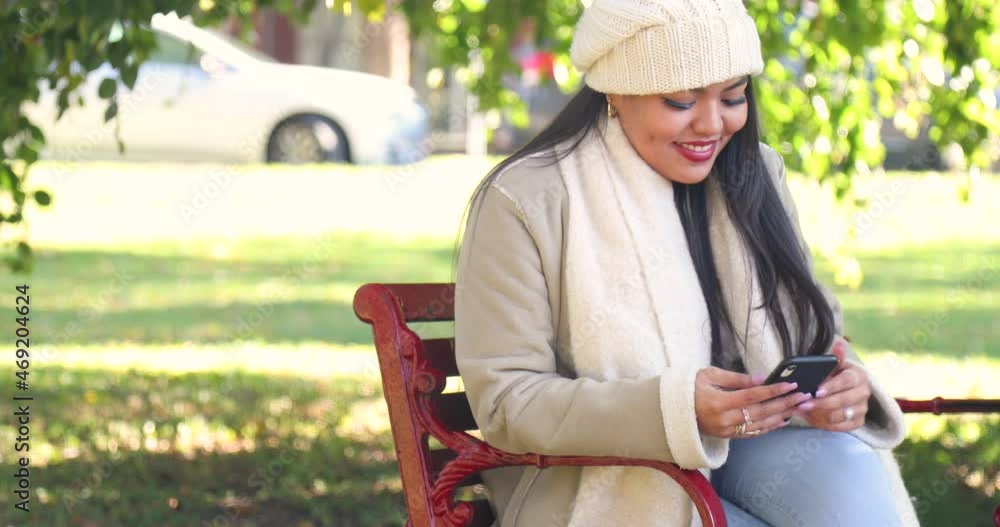 Attractive girl sitting on a city park bench texting during the chilly fall and autumn season. She pauses and looks around and is wearing a white jacket and beanie. Shot in 4k slow motion. Attractive girl sitting on a city park bench texting during the chilly fall and autumn season. She pauses and looks around and is wearing a white jacket and beanie. Shot in 4k slow motion.