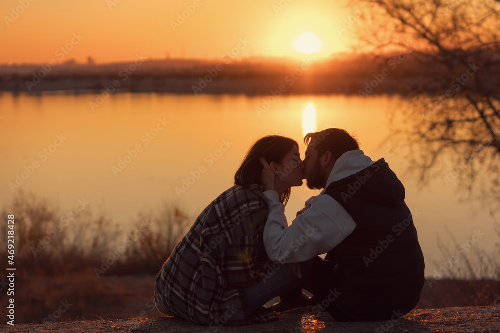 Romantic loving couple kissing while sitting at sunset by lake. Lovers spend time together outdoors. Authentic real people in casual style. Lifestyle Candid portrait. .