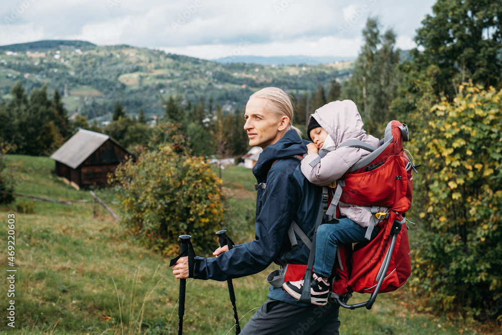 Man traveler with tired child in backpack Father carries his daughter in a backpack Male took slipping kid to the mountains. Traveling with children. Walking with toddler baby in autumn, Nordic walk 