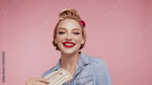 Sexy blond Caucasian woman kiss cash dollars. Girl with red lips hold fan of cash money in dollars banknotes in hands. Female posing isolated on pink background studio with cash. Pop-art, pin up style