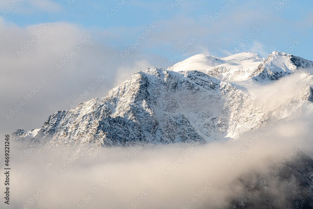 Snow capped mountain peaks in northern Canada with misty, foggy clouds covering the peaks with white, winter theme and spectacular blue sky behind. 