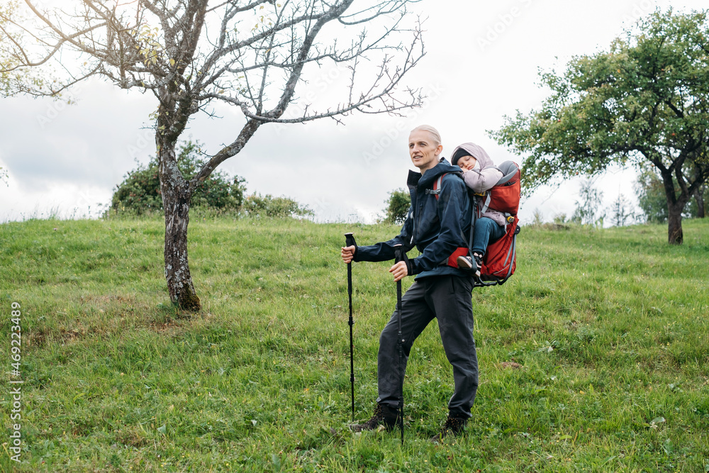 Man hiking with sleepy toddler child in the mountain. Kid sitting in backpack on father back. Active family adventure in forest with tired girl. Cold weather trip, Nordic walk tour Man hiking with sleepy toddler child in the mountain. Kid sitting in backpack on father back. Active family adventure in forest with tired girl. Cold weather trip, Nordic walk tour