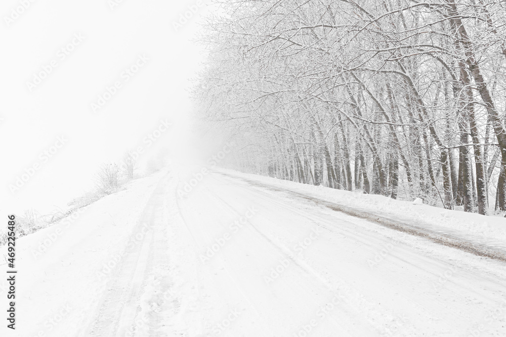 Fototapeta premium Winter road snowfall. An empty rural road without cars, covered with snowdrifts. Travel in bad weather. Winter cloudy landscape with a country road. Tracks of passing cars. The concept of winter magic