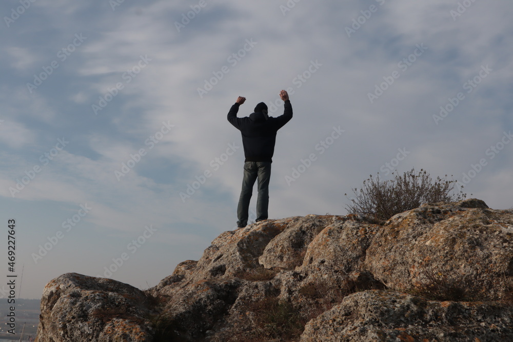 A man with his hands raised stands on a mountain against the blue sky.	