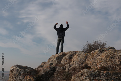 A man with his hands raised stands on a mountain against the blue sky. 