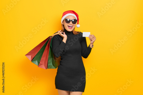 Smiling young Asian woman in Santa Claus hat and sunglasses holding shopping bags and plastic card isolated over yellow background