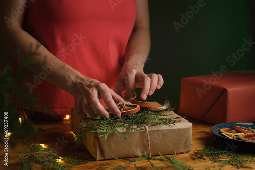 Woman decorates Christmas gift box with dried oranges decoration and Christmas tree twigs. Eco friendly DIY Christmas packaging for holiday gifts.