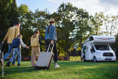 Rear view of young family with suitcases going to caravan outdoors at park.