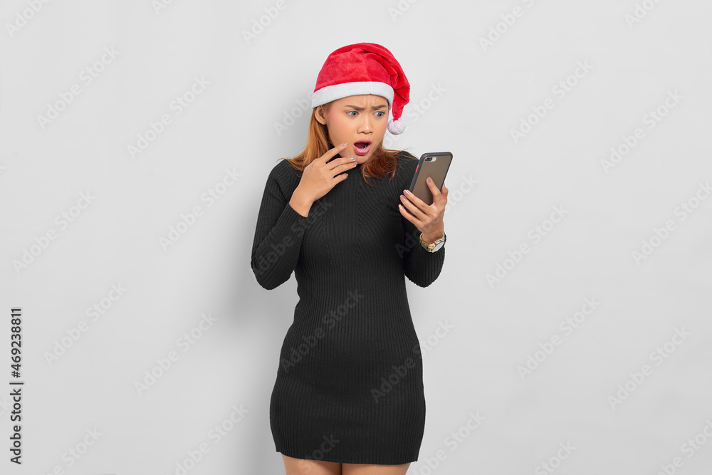 Surprised young Asian woman in Santa Claus hat looking at mobile phone with open mouth isolated over white background Surprised young Asian woman in Santa Claus hat looking at mobile phone with open mouth isolated over white background
