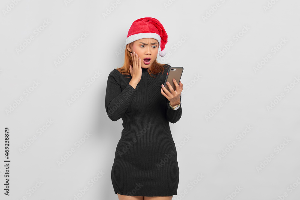 Surprised young Asian woman in Santa Claus hat looking at mobile phone with open mouth isolated over white background Surprised young Asian woman in Santa Claus hat looking at mobile phone with open mouth isolated over white background