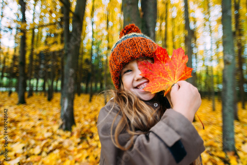 The girl stands in the autumn park and hides her eye behind a maple leaf and smiles.