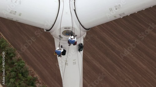 Skilled technician fitters with safety harnesses go down wind turbine pylon blade using long ropes above agricultural plowed field aerial view