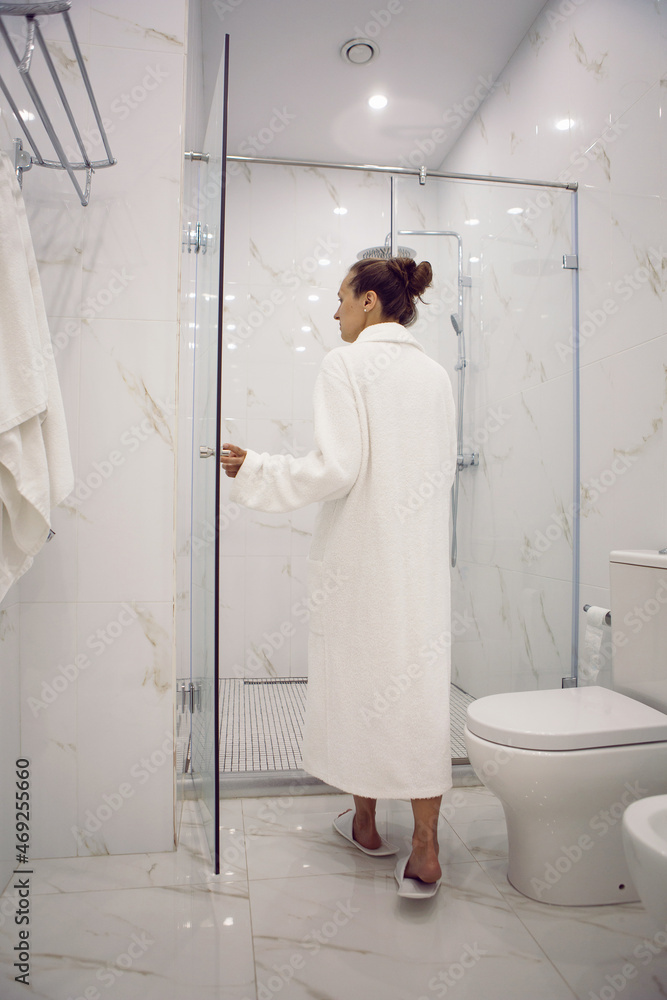 woman standing in the bathroom next to the shower cabin in hotel apartment in a white robe