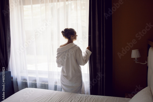 woman in a white robe is stand on a bed by the window in hotel apartment after a shower