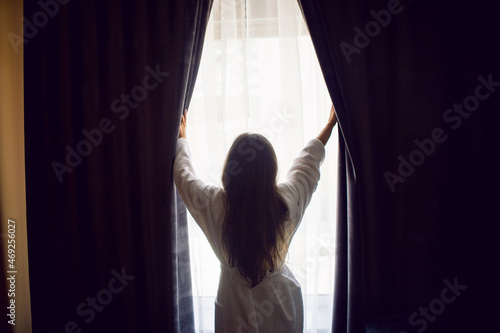 woman stands in a bathrobe at the window and opens the curtain in hotel