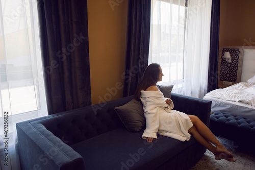 woman in a white robe is sitting on a bed by the window in hotel apartment after a shower