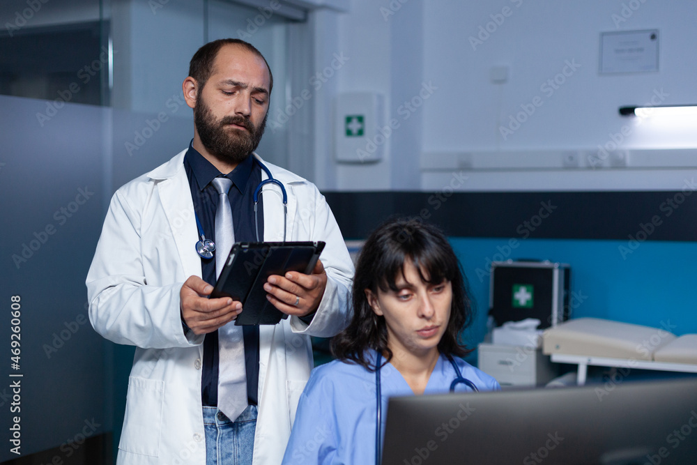 Team of medical workers using technology for overtime work. Doctor holding digital tablet for information while woman nurse looking at computer on desk for healthcare, working late.