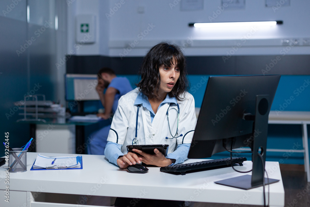 Woman working as doctor with digital tablet and computer for medical information and checkup. Healthcare specialist looking at gadget with touch screen and monitor display at night. Woman working as doctor with digital tablet and computer for medical information and checkup. Healthcare specialist looking at gadget with touch screen and monitor display at night.