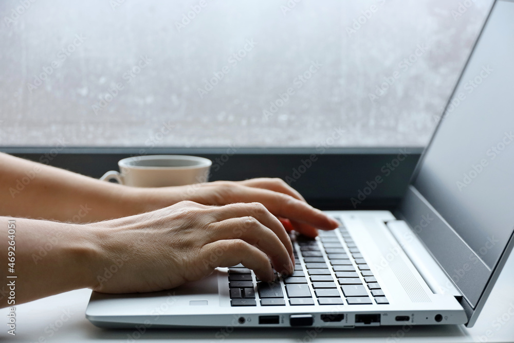 Female hands on laptop keyboard on a table. Woman types on the laptop keyboard sitting near the window Female hands on laptop keyboard on a table. Woman types on the laptop keyboard sitting near the window