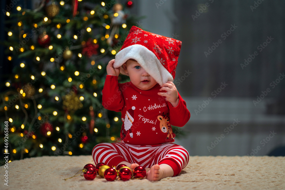 A baby with red hair puts on a santa claus hat against the background of a Christmas tree. Inscription on clothes: Happy New Year