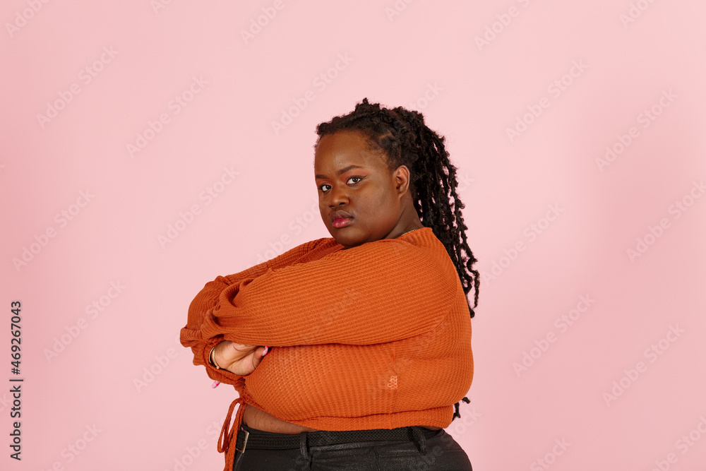 Critical young black plus size body positive woman with dreadlocks in orange top looks at camera crossing arms on pink background in studio Critical young black plus size body positive woman with dreadlocks in orange top looks at camera crossing arms on pink background in studio