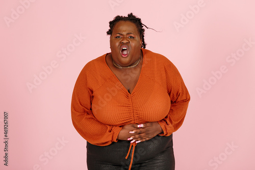 Young black plus size body positive woman patient in orange top suffers from stomach ache standing on light pink background in studio waist up