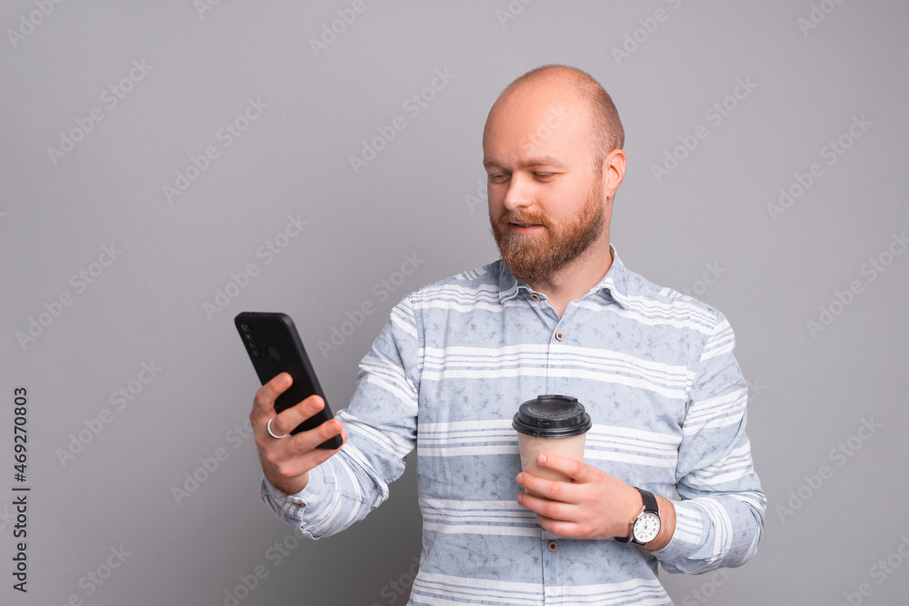 Photo of bearded man standing over grey background and looking at smart phone and holding coffee cup to go. Photo of bearded man standing over grey background and looking at smart phone and holding coffee cup to go.