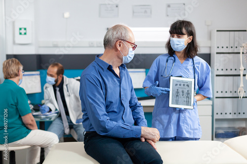 Nurse holding digital tablet with human skeleton on image for elder patient. Medical assistant explaining osteopathy diagnosis with gadget to cure retired man during coronavirus pandemic.
