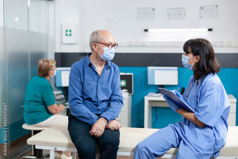 Nurse and patient with face masks doing consultation for osteopathy and healthcare. Medical assistant talking to aged man with physical injury for chiropractic recovery during pandemic Nurse and patient with face masks doing consultation for osteopathy and healthcare. Medical assistant talking to aged man with physical injury for chiropractic recovery during pandemic