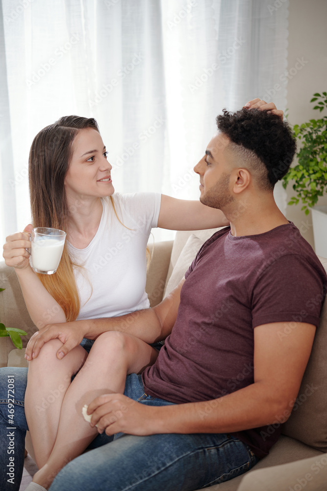 Pretty smiling young woman drinking cup of warm milk and ruffling hair of her husband when they are sitting on sofa Pretty smiling young woman drinking cup of warm milk and ruffling hair of her husband when they are sitting on sofa